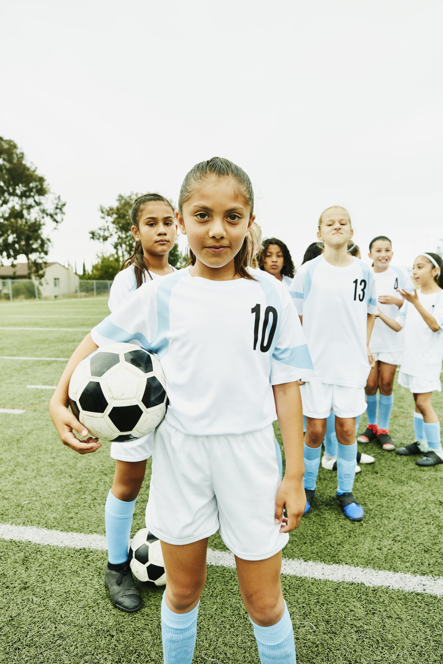 Portrait of young female soccer player holding ball and standing in front of teammates