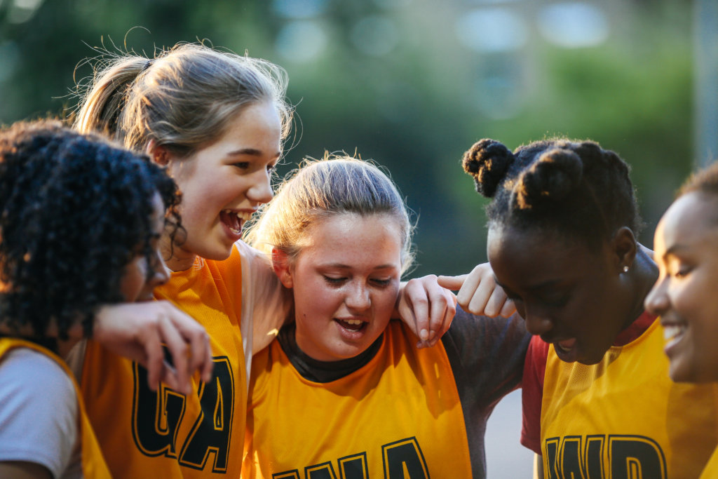 team of young netball players, in huddle with arms around each other on outdoor netball court